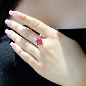 May include: A gold ring with a floral design, set with bright red gemstones. The ring is worn on a hand with manicured nails, against a dark backdrop. The gemstones are a deep red colour.
