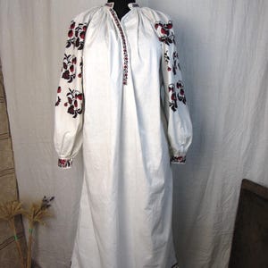 Puede incluir: Un vestido blanco de manga larga con escote dividido y hombros fruncidos. La prenda está decorada con intrincados bordados florales en rojo y negro a lo largo de las mangas, los puños, el escote y el dobladillo inferior del vestido.