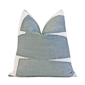 May include: A square decorative pillow with a blue-grey background and white geometric shapes. The pillow has a textured appearance and a soft, inviting look. The design is modern and abstract, suitable for home decor.