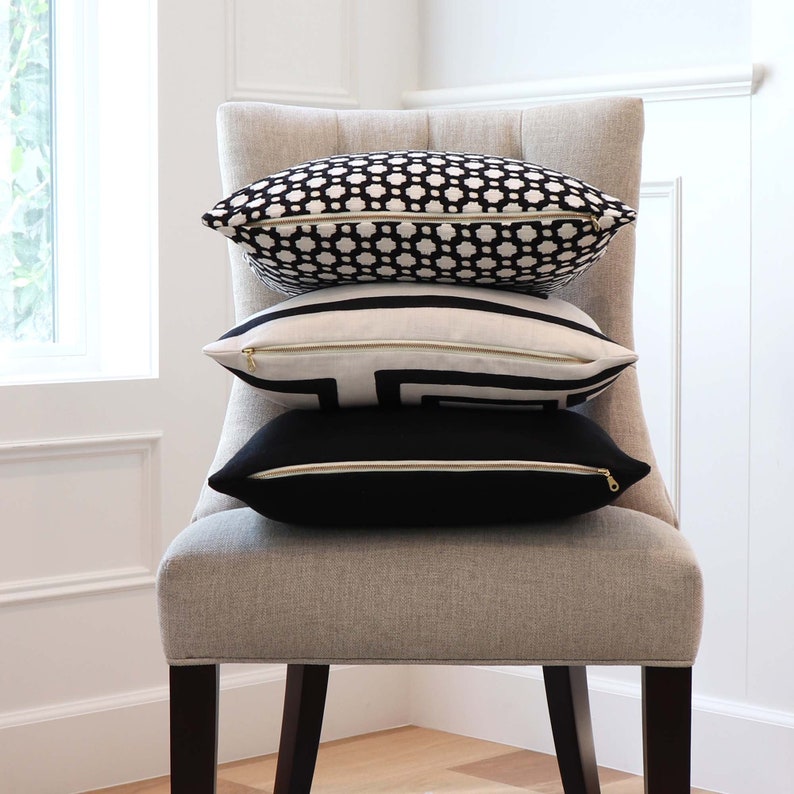 May include: A stack of three decorative throw pillows on a light grey chair. The top pillow has a black and white geometric pattern, the middle pillow has a black and white geometric pattern, and the bottom pillow is solid black.
