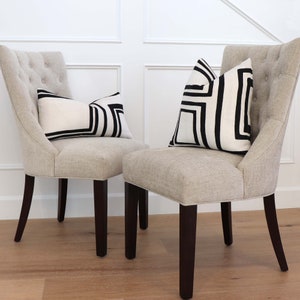 May include: Two beige upholstered chairs with dark brown legs. Each chair has a black and white geometric patterned cushion on the seat.