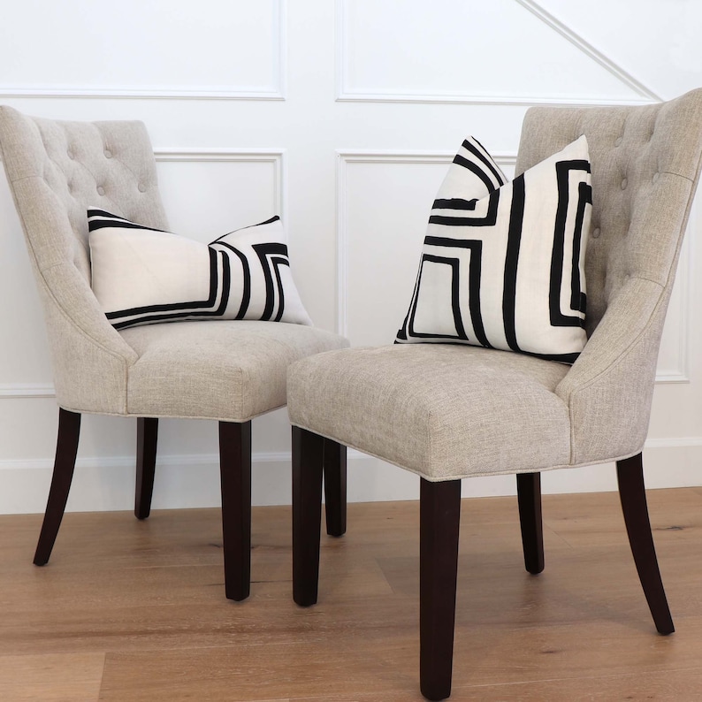 May include: Two beige upholstered chairs with dark brown legs. Each chair has a black and white geometric patterned cushion on the seat.