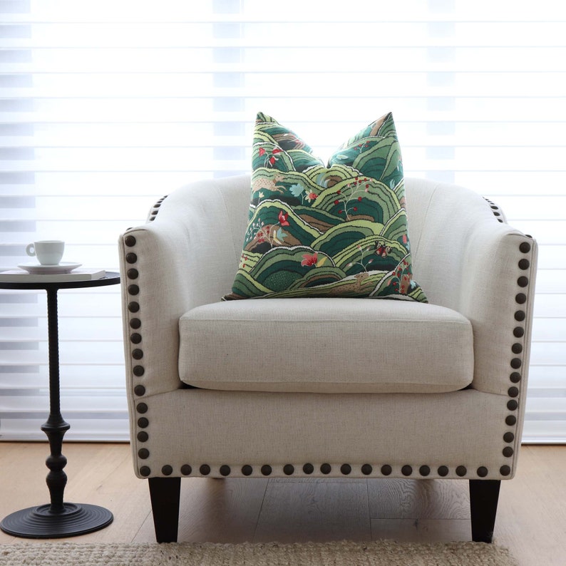 May include: A white upholstered armchair with a green and brown patterned pillow. The armchair has a curved back and a tufted seat. The armchair is accented with dark brown nailhead trim.