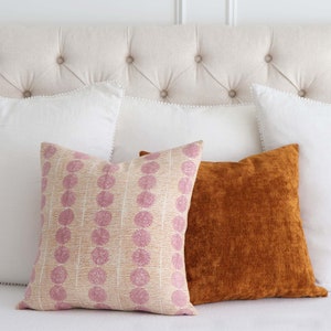 May include: Two decorative throw pillows on a white bed. One pillow has a pink and white geometric pattern, and the other is a solid rust orange.
