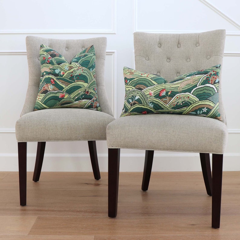 May include: Two beige chairs with dark brown legs and green and red patterned throw pillows. The pillows have a landscape design with a stylized mountain range and a small animal.