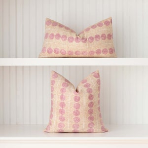 May include: Two decorative throw pillows with a pink and beige geometric pattern. The pillows are on a white shelf.