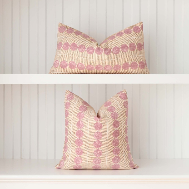 May include: Two decorative throw pillows with a pink and beige geometric pattern. The pillows are on a white shelf.