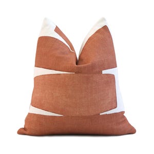 May include: A decorative throw pillow with a terracotta-colored background and white geometric shapes. The pillow has a soft, textured appearance and is designed for home decor. The pillow is square-shaped.