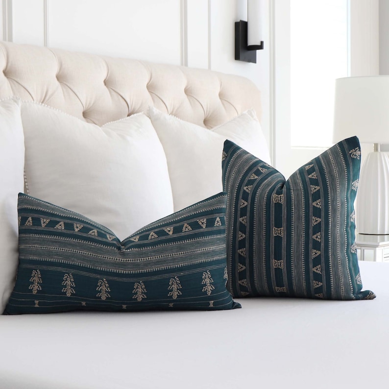 Puede incluir: Dos almohadas decorativas con un patr&oacute;n geom&eacute;trico azul verdoso y dorado sobre una cama blanca.