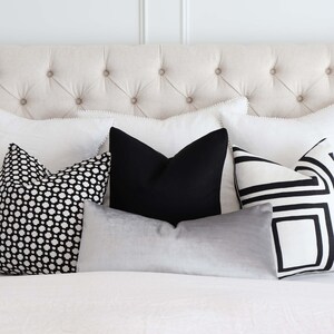 May include: A white bed with a tufted headboard and a variety of black and white patterned throw pillows. The pillows are arranged in a row on the bed, with a grey velvet pillow in the front.