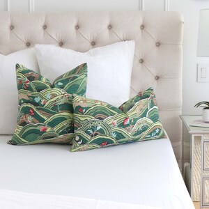 May include: Two green and brown patterned throw pillows with a white background and a white bedspread. The pillows feature a landscape design with hills, trees, and flowers.