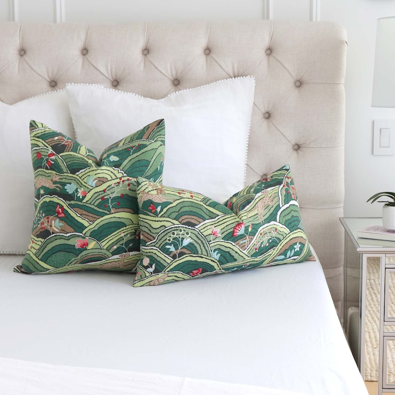 May include: Two green and brown patterned throw pillows with a white background and a white bedspread. The pillows feature a landscape design with hills, trees, and flowers.