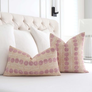 May include: Two decorative throw pillows with a pink and cream geometric pattern. The pillows are on a white bed with white bedding.