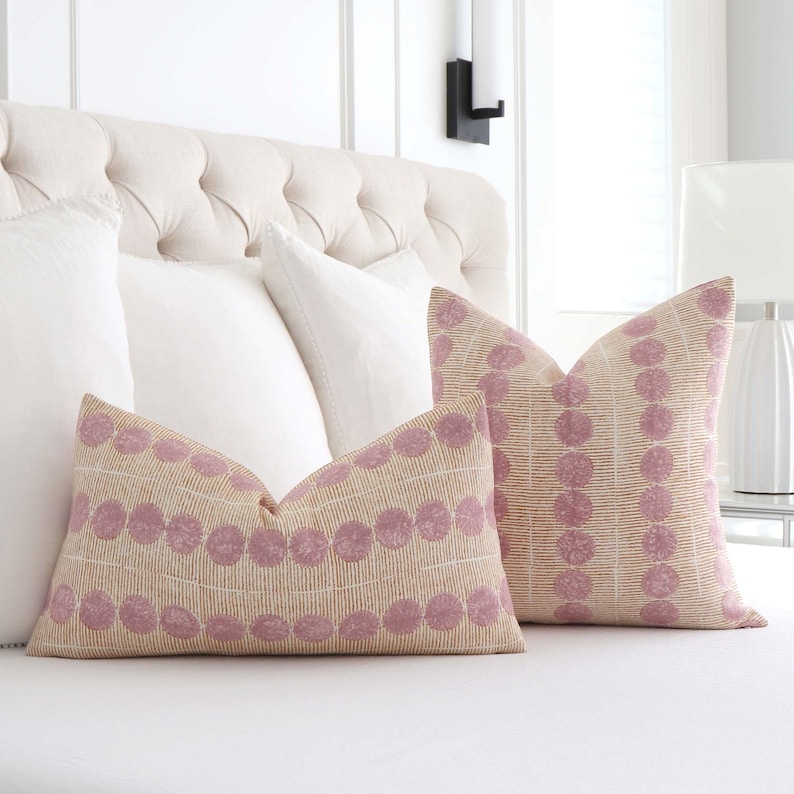 May include: Two decorative throw pillows with a pink and cream geometric pattern. The pillows are on a white bed with white bedding.