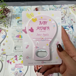 May include: Pink baby shower bottle opener favours with a hot air balloon design. The opener features the text "Baby Shower" and the name "Jane Smith". The favours are decorated with baby-themed graphics and the text "Thank you for celebrating with me!"