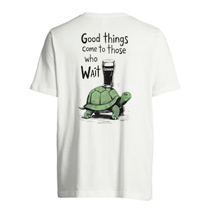 Pode incluir: Camiseta branca com um desenho de tartaruga verde carregando um copo de Guinness. O texto diz "Coisas boas vêm para aqueles que esperam". O copo de Guinness é preto com texto branco. A tartaruga é verde e marrom.