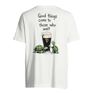 Pode incluir: Camiseta branca com uma ilustração de desenho animado de duas tartarugas verdes e uma caneca de cerveja preta com espuma. O texto "Good things come to those who wait" está impresso acima da ilustração.