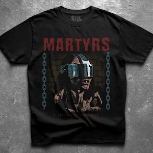 May include: A black short-sleeved t-shirt featuring a graphic print with the red text MARTYRS at the top. The design includes a central figure wearing a metallic visor, flanked by two vertical blue chains against a dark brick wall background.