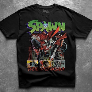 Könnte beinhalten: Ein schwarzes Kurzarm-T-Shirt mit einer Grafik der Comicfigur Spawn mit rotem Umhang und Ketten. Das Wort SPAWN ist in Grün über der Figur gedruckt und der Text TODD MCFARLANE ist in Rot am unteren Rand des T-Shirts zu sehen.
