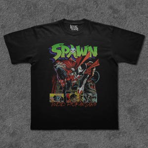 May include: Black t-shirt featuring a comic book illustration of Spawn, a superhero character. The design includes the word "Spawn" in green and purple, a skull, and the artist's name, Todd McFarlane. The shirt has a crew neck and short sleeves.