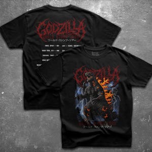 May include: A black short-sleeve t-shirt featuring a graphic of Godzilla with orange glowing accents against a blue lightning background. The back displays the text Godzilla World Stomp Tour with a list of cities and years in red lettering.