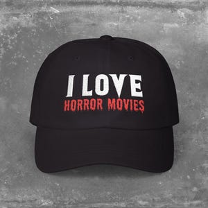Puede incluir: Gorra de béisbol negra con el texto "I LOVE HORROR MOVIES" en letras blancas y rojas. La gorra tiene una visera curva y una corona estructurada. El texto está centrado en la parte delantera.