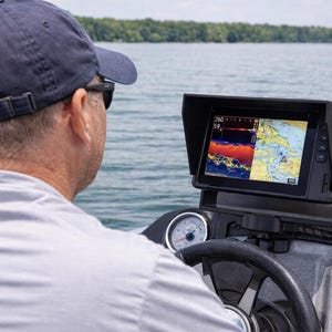 May include: A person navigating a boat, looking at a marine navigation system. The screen shows sonar and map data. The boat's dashboard includes gauges and a steering wheel. The background shows a lake and trees.