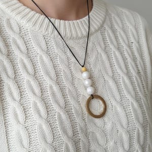 May include: A necklace with a black cord, featuring a wooden bead, three white beads, and a large gold-coloured ring pendant. The necklace is displayed against a white cable-knit jumper.