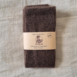 May include: A pair of brown baby leg warmers made from 100% organic sheep wool. The product is wrapped with a tan paper band that reads "Baby Leg Warmers" and "0-3 years". The brand name "Crunchy Cult" and "Made in Mongolia" are also printed on the band.