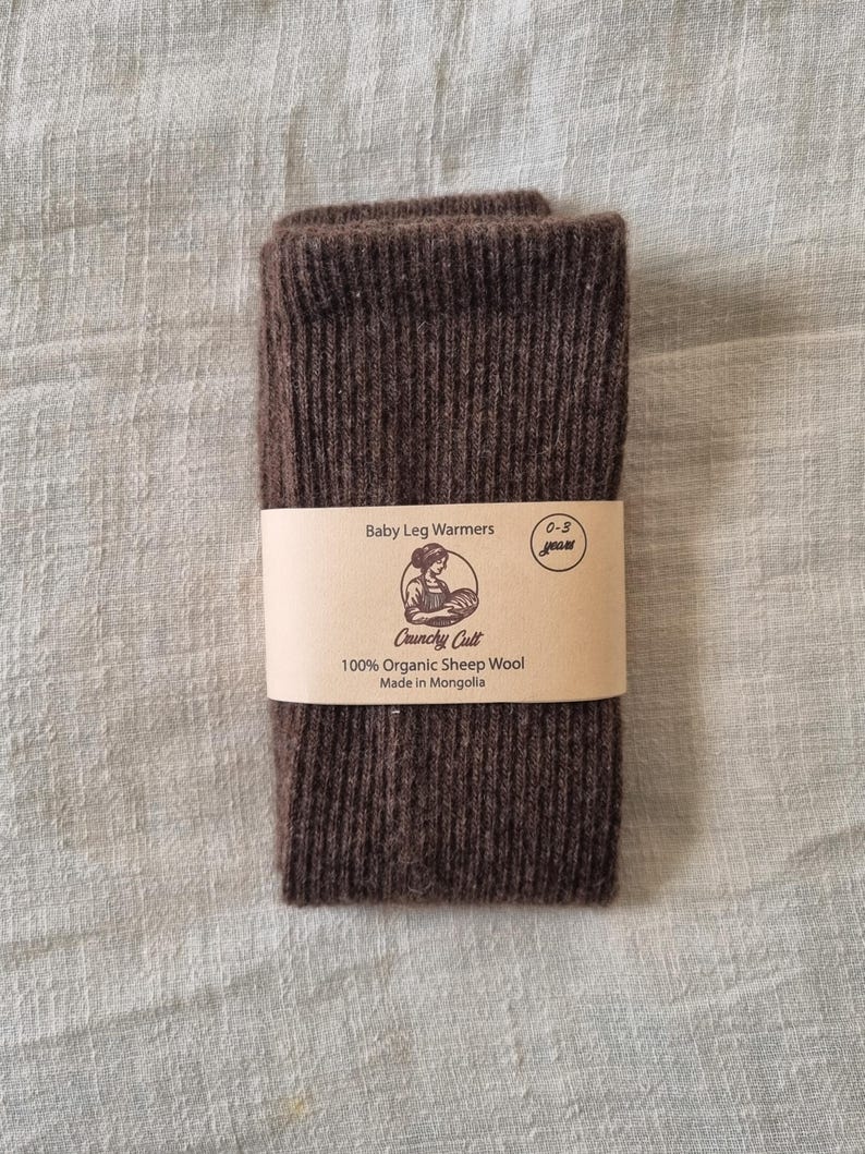 May include: A pair of brown baby leg warmers made from 100% organic sheep wool. The product is wrapped with a tan paper band that reads "Baby Leg Warmers" and "0-3 years". The brand name "Crunchy Cult" and "Made in Mongolia" are also printed on the band.