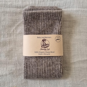 May include: Folded pair of gray baby leg warmers with a ribbed knit texture. A brown paper band wraps around the warmers, displaying the text "Baby Leg Warmers," "Crunchy Cult," and "100% Organic Sheep Wool." The label also indicates the product is made in Mongolia.