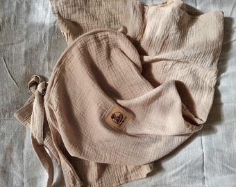 Puede incluir: Un juego de manta y gorro para bebé de muselina beige. La manta está doblada y drapeada, con un aspecto texturizado. Una pequeña etiqueta cuadrada con un logotipo es visible en el gorro. El conjunto incluye lazos largos para asegurar la manta.