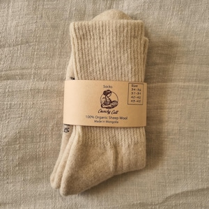 Könnte beinhalten: Ein Paar beige Socken, gefaltet und mit einem braunen Papierband umwickelt. Auf dem Band steht "Socks", "Crunchy Cult", "100% Organic Sheep Wool" und "Made in Mongolia". Größenangaben sind ebenfalls aufgedruckt.