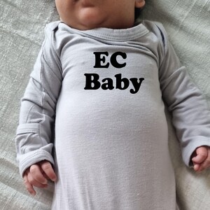 May include: A light gray baby onesie with long sleeves. The text "EC Baby" is printed in bold black letters on the front. The baby is lying on a light gray surface.