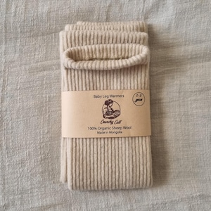 May include: A pair of beige baby leg warmers made of 100% organic sheep wool. The leg warmers are folded and wrapped with a brown paper band that reads "Baby Leg Warmers" and "0-3 years". The label also includes the text "Crunchy Cult" and "Made in Mongolia".