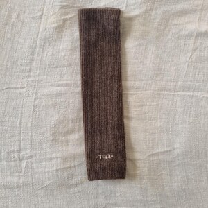 May include: A brown knitted arm warmer with the text "-TOA-" embroidered in white near the cuff. The arm warmer is rectangular and made of a ribbed knit fabric. The background is a white, textured fabric.