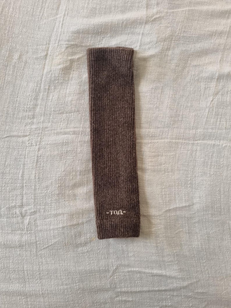 May include: A brown knitted arm warmer with the text "-TOA-" embroidered in white near the cuff. The arm warmer is rectangular and made of a ribbed knit fabric. The background is a white, textured fabric.