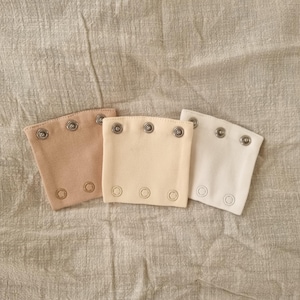 Puede incluir: Tres piezas de tela en tonos beige, crema y blanco, cada una con cierres de broche metálicos. Están dispuestas sobre un fondo de tela texturizada de tono neutro. Las piezas de tela tienen forma rectangular con esquinas redondeadas.