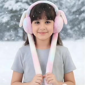 May include: A child wearing fluffy, pastel-coloured earmuffs with long, dangling bunny ears. The earmuffs are primarily pink, with hints of blue and yellow, set against a snowy background. The child is smiling.