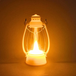 Puede incluir: Una linterna blanca que funciona con pilas con una carcasa de vidrio transparente y una llama LED brillante y parpadeante. La linterna tiene un asa y una parte superior decorativa, creando un brillo cálido. La linterna está diseñada para parecerse a una lámpara de aceite tradicional.