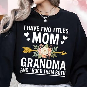 May include: Black sweatshirt with the text "I HAVE TWO TITLES MOM GRANDMA AND I ROCK THEM BOTH". The design includes a floral arrow and heart accents. A casual top for mothers and grandmothers.
