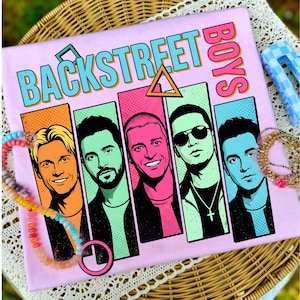 Pode incluir: T-shirt rosa claro com a inscrição "BACKSTREET BOYS" em turquesa e rosa. A camisa apresenta um gráfico de cinco homens em painéis coloridos. Uma pulseira de contas e um grampo de cabelo são visíveis.