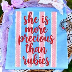 Puede incluir: Una tela azul claro con un diseño de rayas verticales rosas y blancas y el texto "she is more precious than rubies" en escritura roja. La tela está decorada con un lazo rosa y una colección de pulseras.