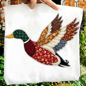 May include: A white fabric item featuring a colorful appliqué of a flying duck. The duck has a green plaid head, a red floral patterned body, and wings with brown, tan, and blue patterned details. The duck's beak is yellow.