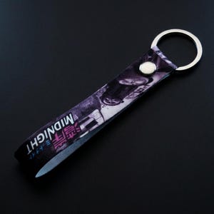 May include: A black and purple keychain featuring a car graphic and the word "MIDNIGHT" in white and pink. The keychain has a silver metal ring and a silver press stud.