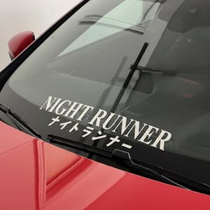 May include: White "NIGHT RUNNER" decal with Japanese characters on a car windshield. The decal is centered and positioned above the windshield wipers. The car's hood is red.