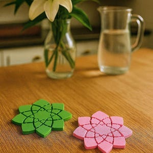 May include: A wooden table displays two flower-shaped coasters, one green and one pink. A vase of white lilies and a glass pitcher are in the background. The coasters have a detailed, geometric design.