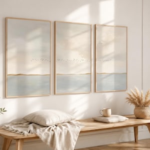 Op de afbeelding: Een set van drie verticale wandkunstprints in lichte houten lijsten, elk met een sereen landschap met een lichtblauwe waterhorizon, een dunne gouden lijn, een zachte beige lucht en een kleine zwerm zwarte vogels.