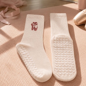Op de afbeelding: Een paar lichtbeige enkelsokken met een kleine geborduurde afbeelding van donkerroze balletschoenen op de buitenkant van de enkel. De zolen van de sokken zijn voorzien van een textuurpatroon van kleine, verhoogde witte siliconen hartjes.