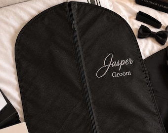 May include: A black garment bag with a central zipper closure. The front features the white embroidered text "Jasper" in a script font above the word "Groom" in a sans-serif font. The bag is suspended by a metal hanger hook at the top.